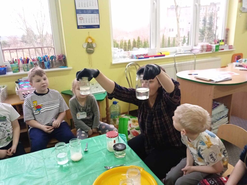 Zdęcie przedstawia panią oraz dzieci siedzące wokół stolika w sali przedszkolnej. Pani w rękach trzyma pojemniki z wodą.