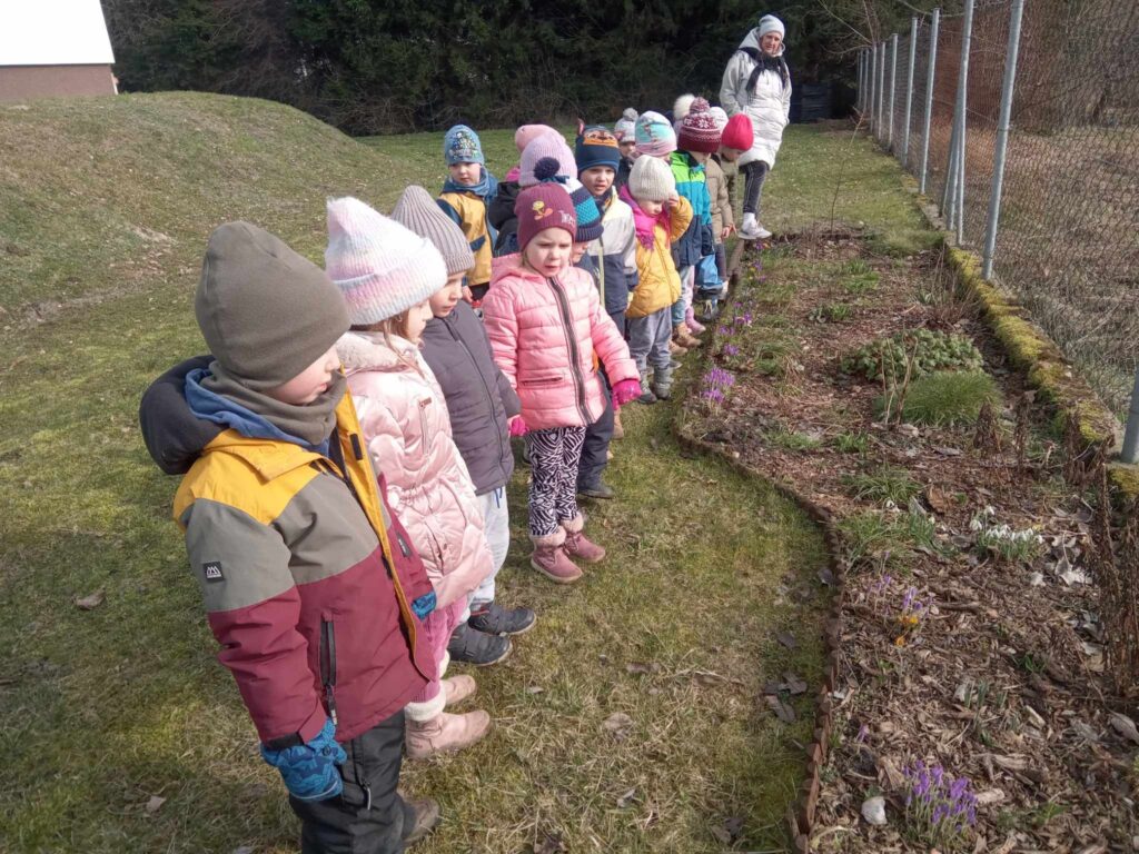 Zdjęcie przedstawia panią oraz dzieci poszukujące oznak wiosny w ogrodzie przedszkolnym.