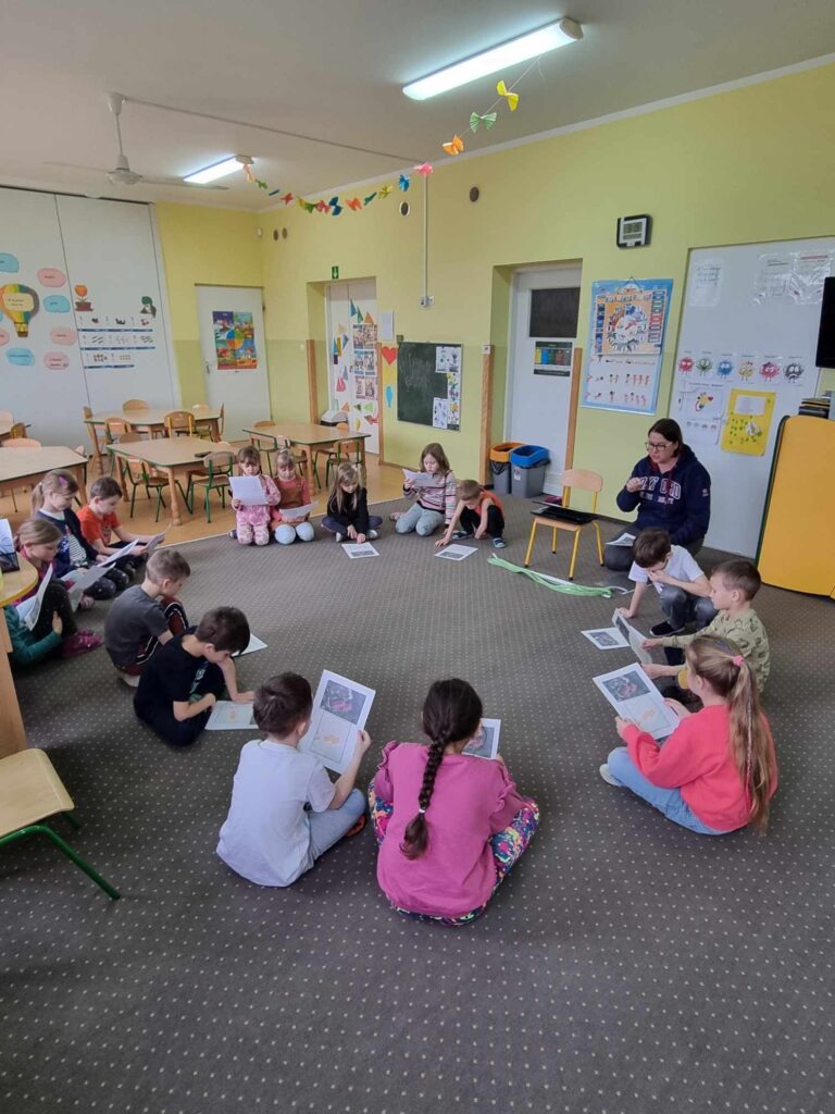 Zdjęcie przedstawia panią oraz dzieci siedzące na wykładzinie w kole w sali przedszkolnej.