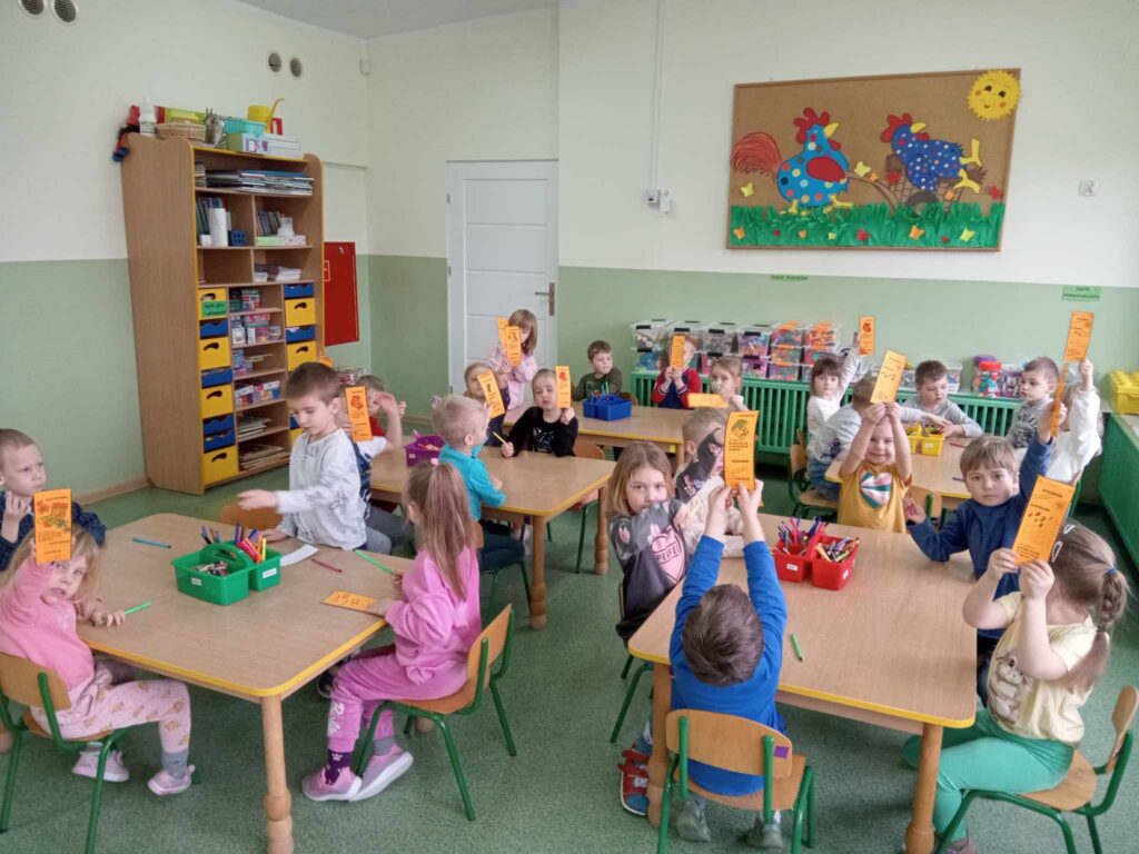 Zdjęcie przedstawia dzieci siedzące przy stolikach w sali przedszkolnej. Wykonują prace plastyczne.