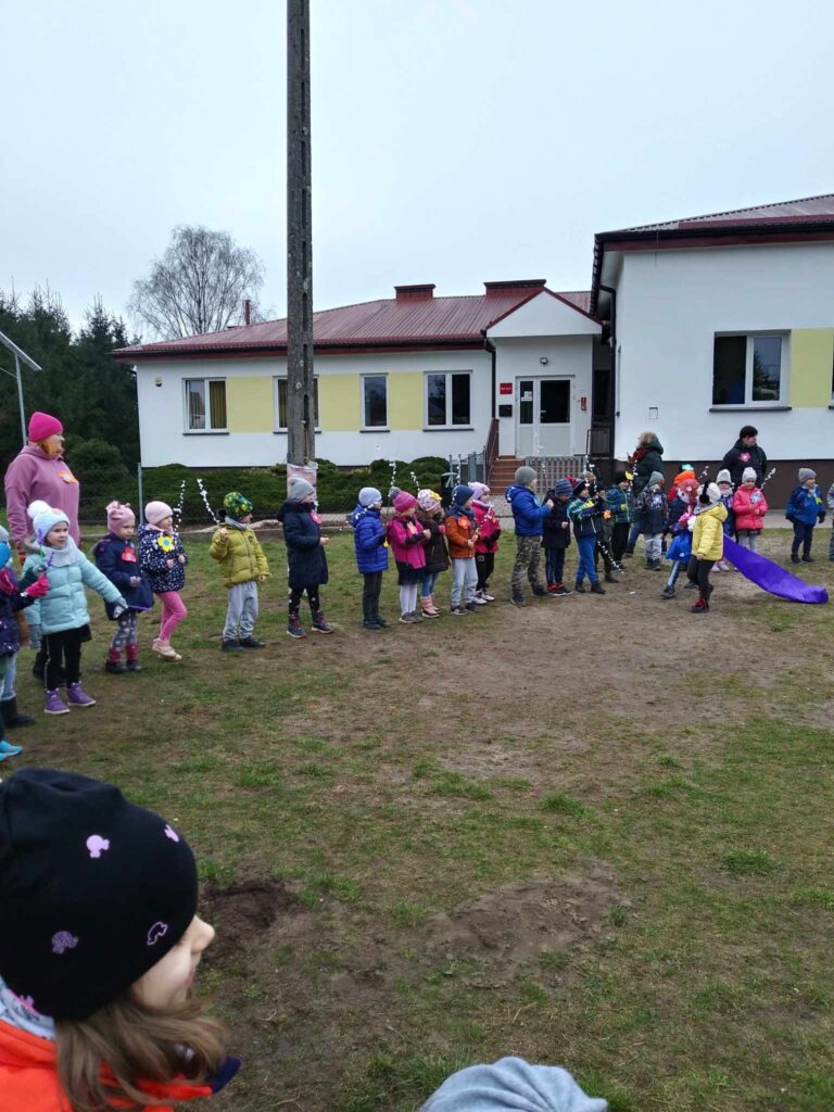 Zdjęcie przedstawia panie oraz dzieci - wszyscy witają wiosnę na placu przedszkolnym.