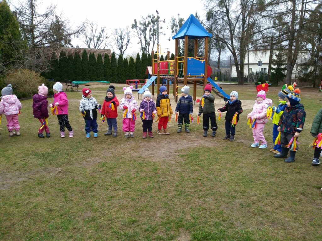 Zdjęcie przedstawia dzieci - wszyscy witają wiosnę na placu przedszkolnym.