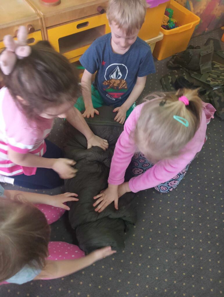 Zdjęcie przedstawia dzieci siedzące na dywanie w sali przedszkolnej - składają koc.