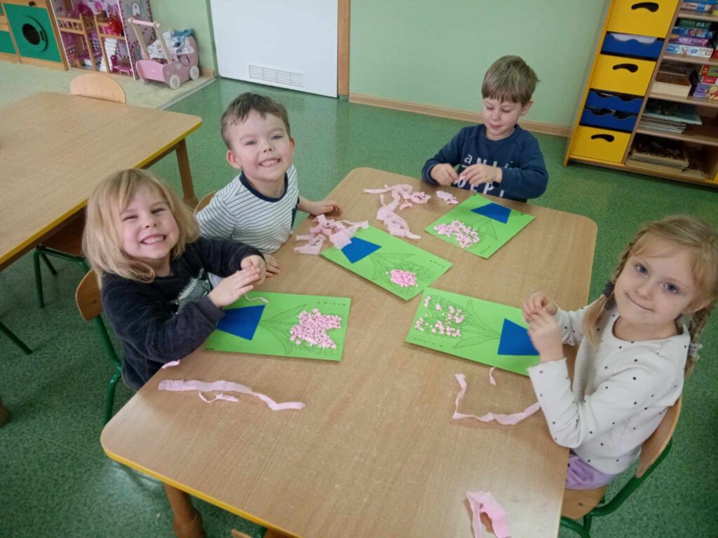 Zdjęcie przedstawia dzieci siedzące przy stoliku w sali przedszkolnej. Wykonują prace plastyczne.