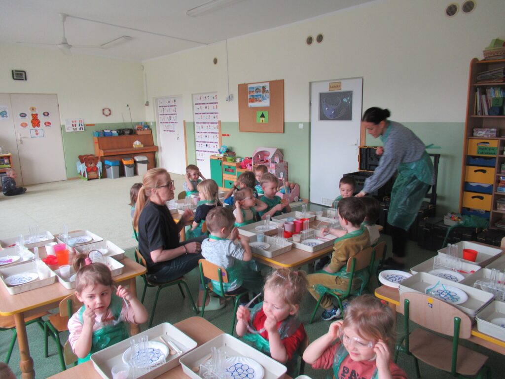 Zdjęcie przedstawia panie oraz siedzące dzieci wokół stolików w sali przedszkolnej. Dzieci wykonują doświadczenia.