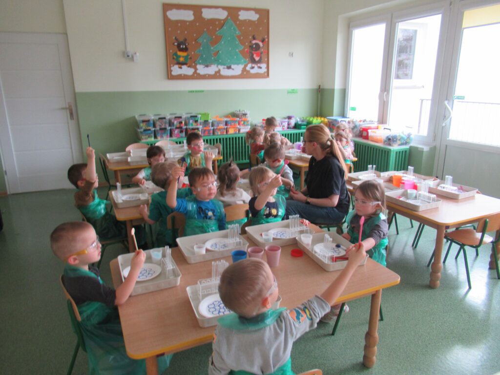 Zdjęcie przedstawia panią oraz siedzące dzieci wokół stolików w sali przedszkolnej. Dzieci wykonują doświadczenia.