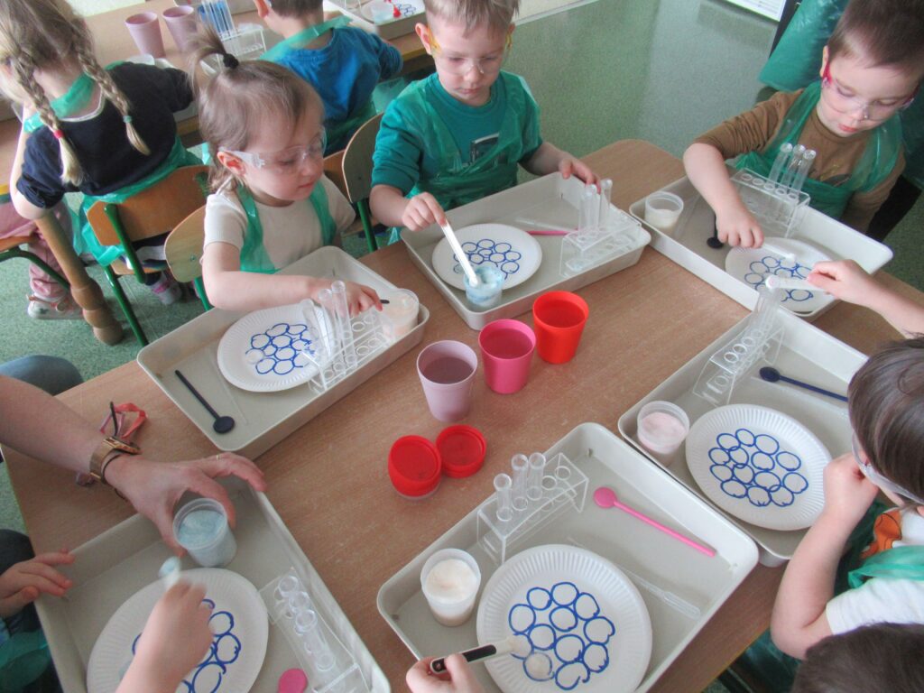 Zdjęcie przedstawia siedzące dzieci wokół stolików w sali przedszkolnej. Dzieci wykonują doświadczenia.