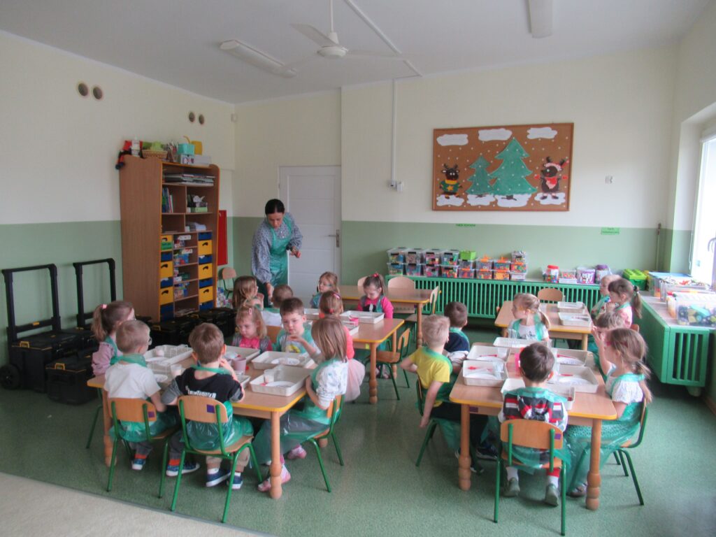Zdjęcie przedstawia panią oraz siedzące dzieci wokół stolików w sali przedszkolnej. Dzieci wykonują doświadczenia.