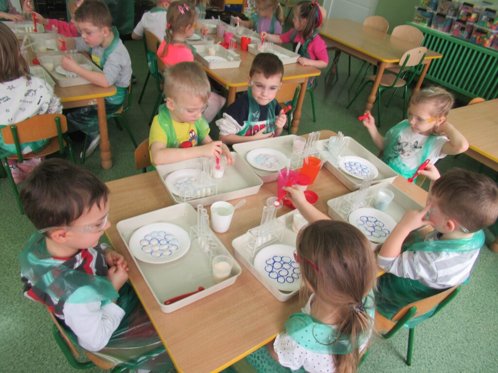 Zdjęcie przedstawia siedzące dzieci wokół stolików w sali przedszkolnej. Dzieci wykonują doświadczenia.