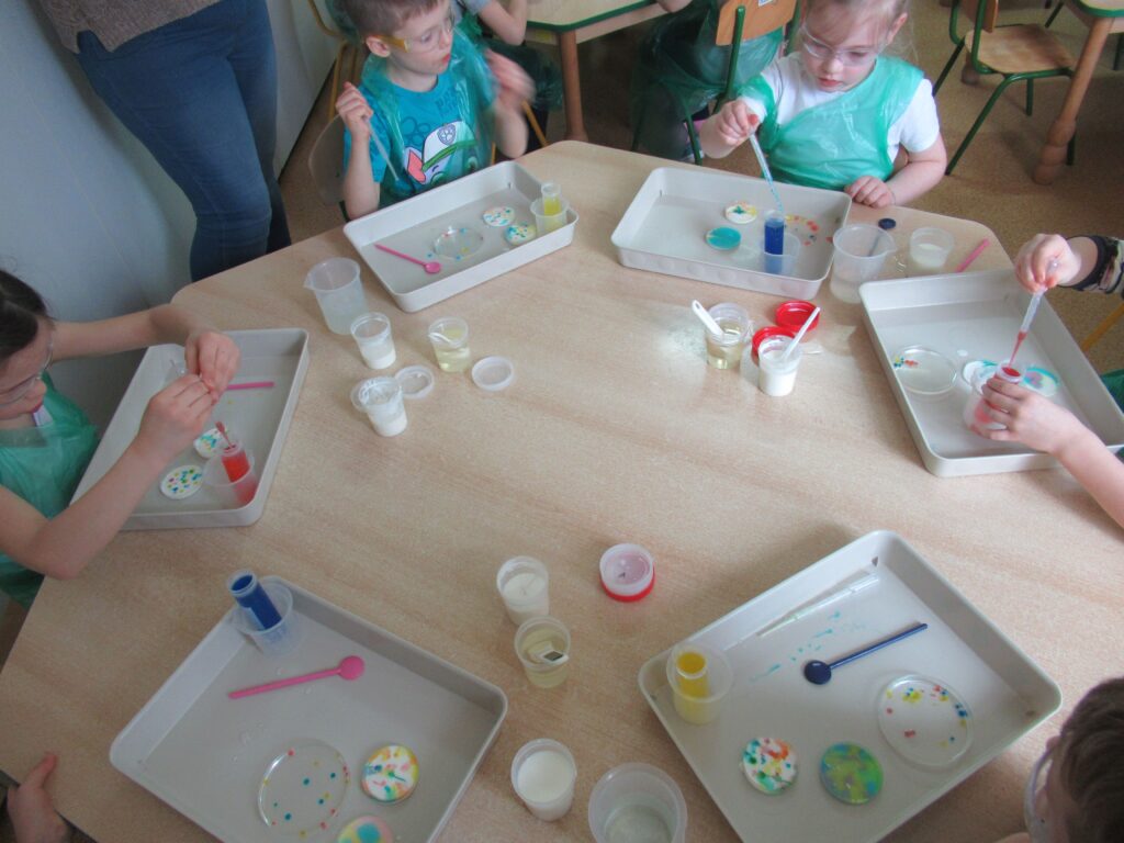 Zdjęcie przedstawia dzieci siedzące przy stoliku w sali przedszkolnej. Na stoliku stoją różne pojemniczki i inne akcesoria do robienia doświadczeń.