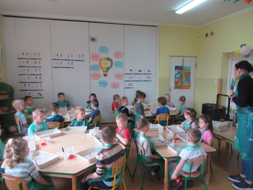 Zdjęcie przedstawia panią oraz dzieci siedzących wokół stolików w sali przedszkolnej. Dzieci wykonują doświadczenia.