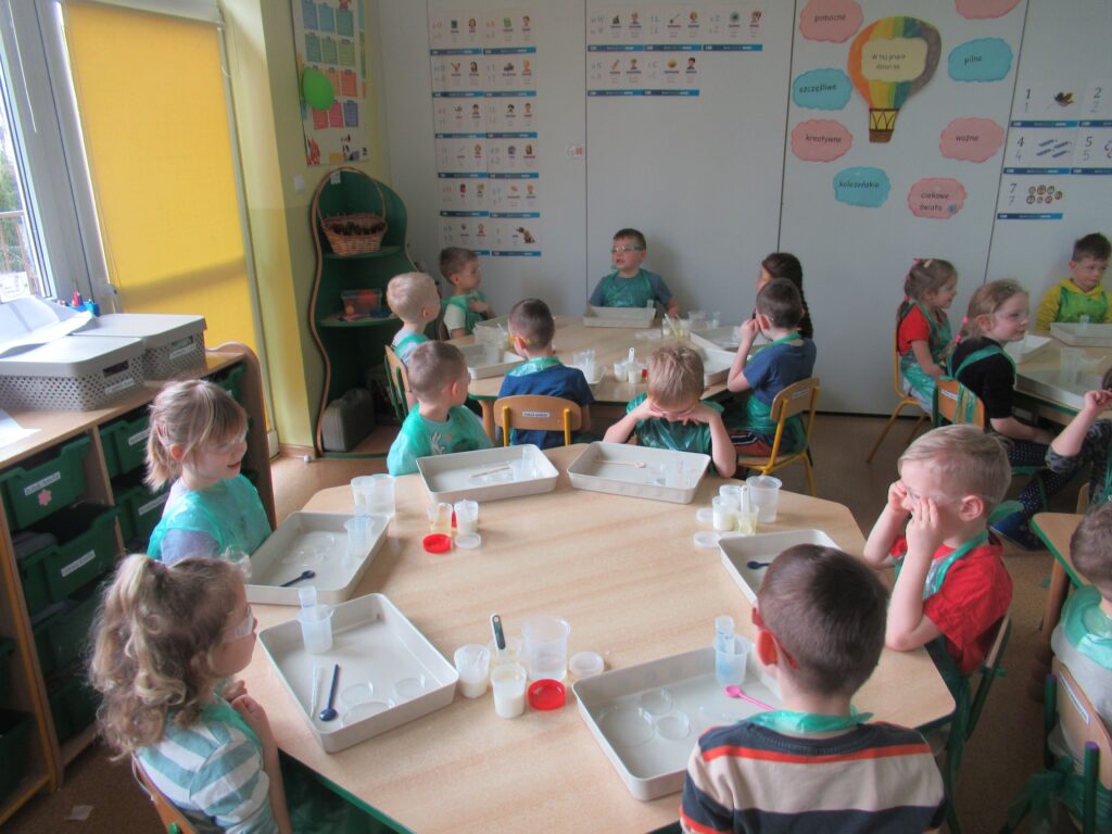 Zdjęcie przedstawia dzieci siedzących wokół stolików w sali przedszkolnej. Dzieci wykonują doświadczenia.