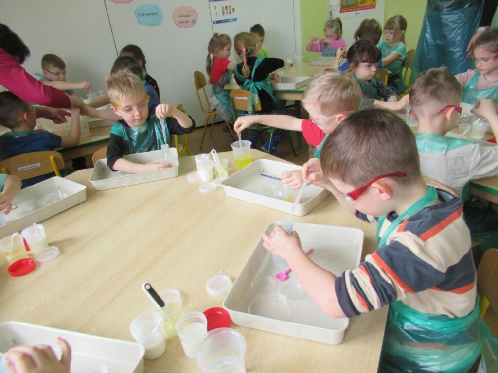 Zdjęcie przedstawia dzieci siedzących wokół stolików w sali przedszkolnej. Dzieci wykonują doświadczenia.