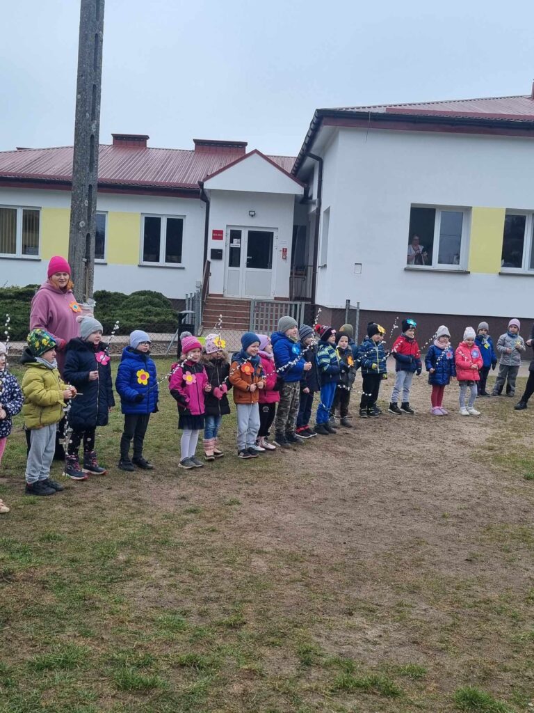 Zdjęcie przedstawia panią oraz dzieci - wszyscy witają wiosnę na placu przedszkolnym.