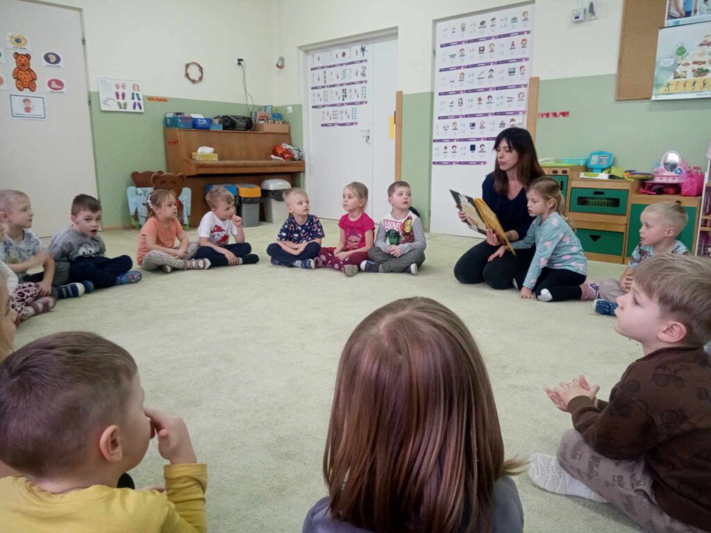 Zdjęcie przedstawia panią oraz dzieci siedzące na wykładzinie w kole w sali przedszkolnej.