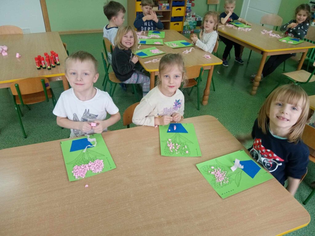Zdjęcie przedstawia dzieci siedzące przy stolikach w sali przedszkolnej. Wykonują prace plastyczne.