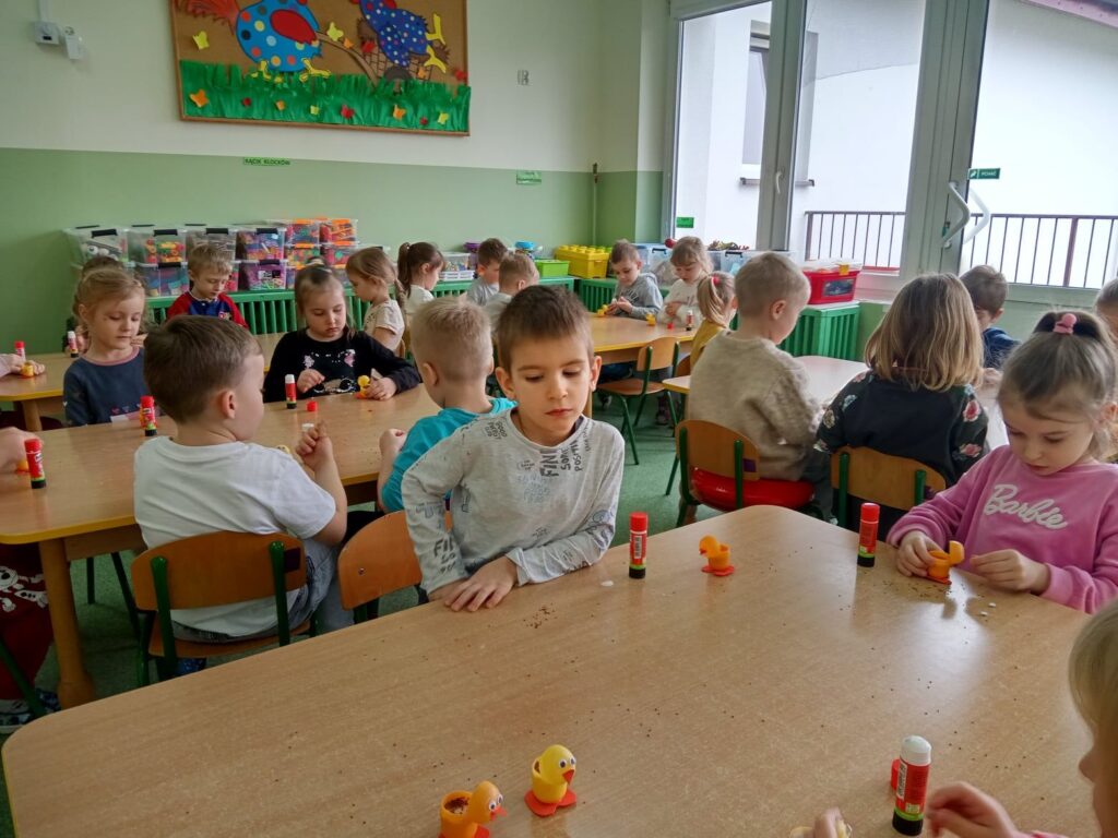 Zdjęcie przedstawia dzieci siedzące przy stolikach na zajęciach w sali przedszkolnej.