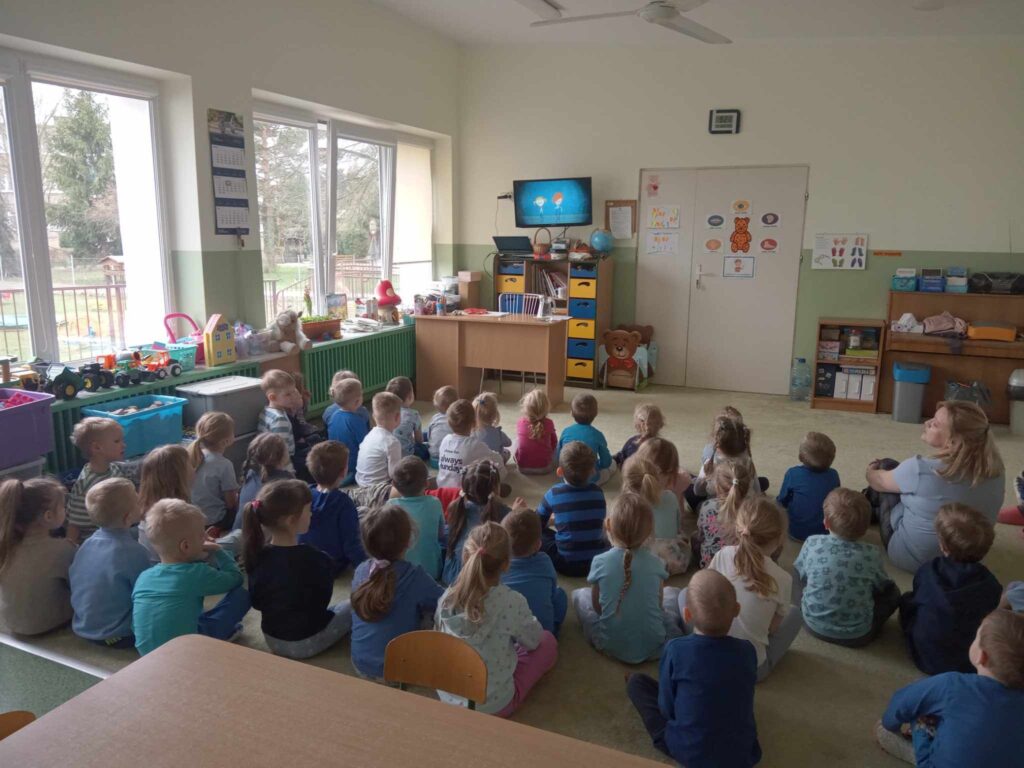 Zdjęcie przedstawia panią oraz dzieci w sali przedszkolnej.