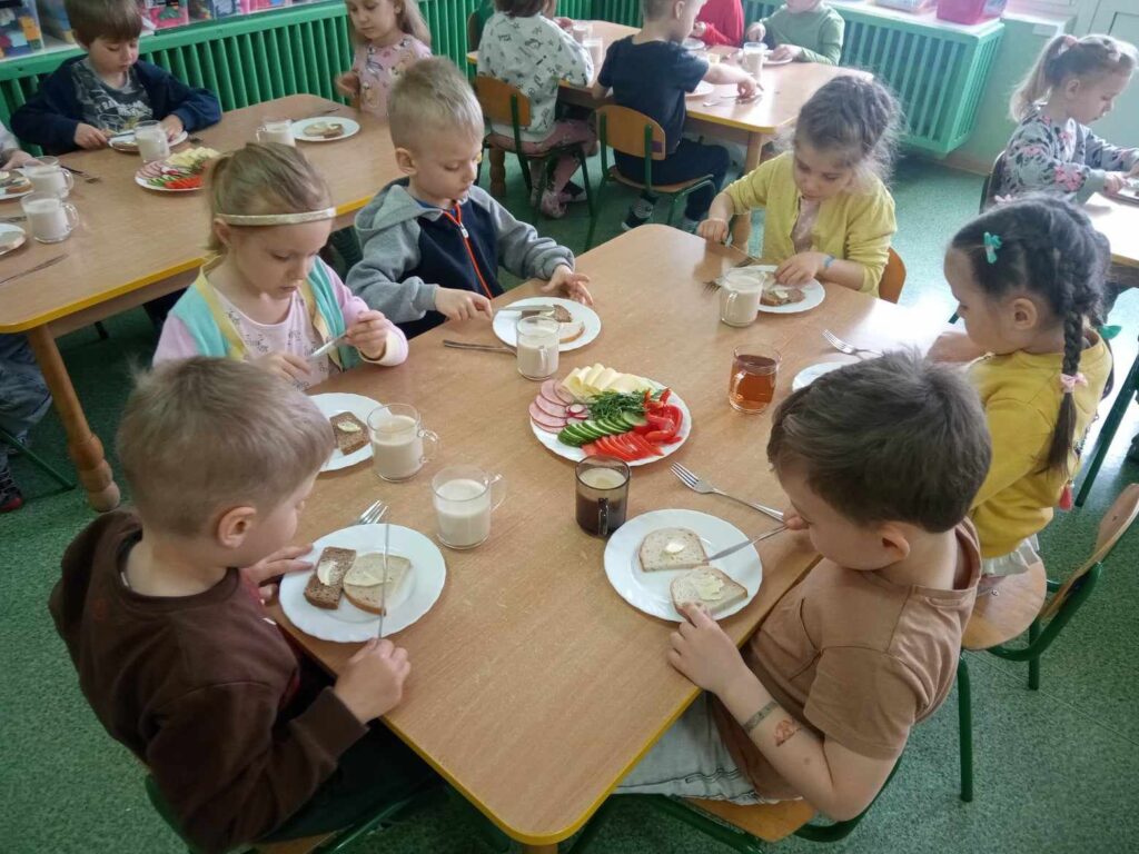 Zdjęcie przedstawia dzieci siedzące przy stolikach w sali przedszkolnej.