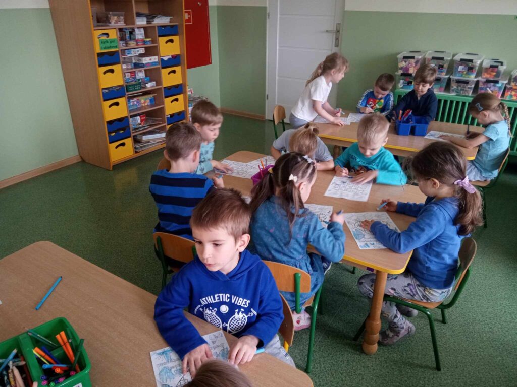 Zdjęcie przedstawia dzieci siedzące w sali przedszkolnej przy stolikach.