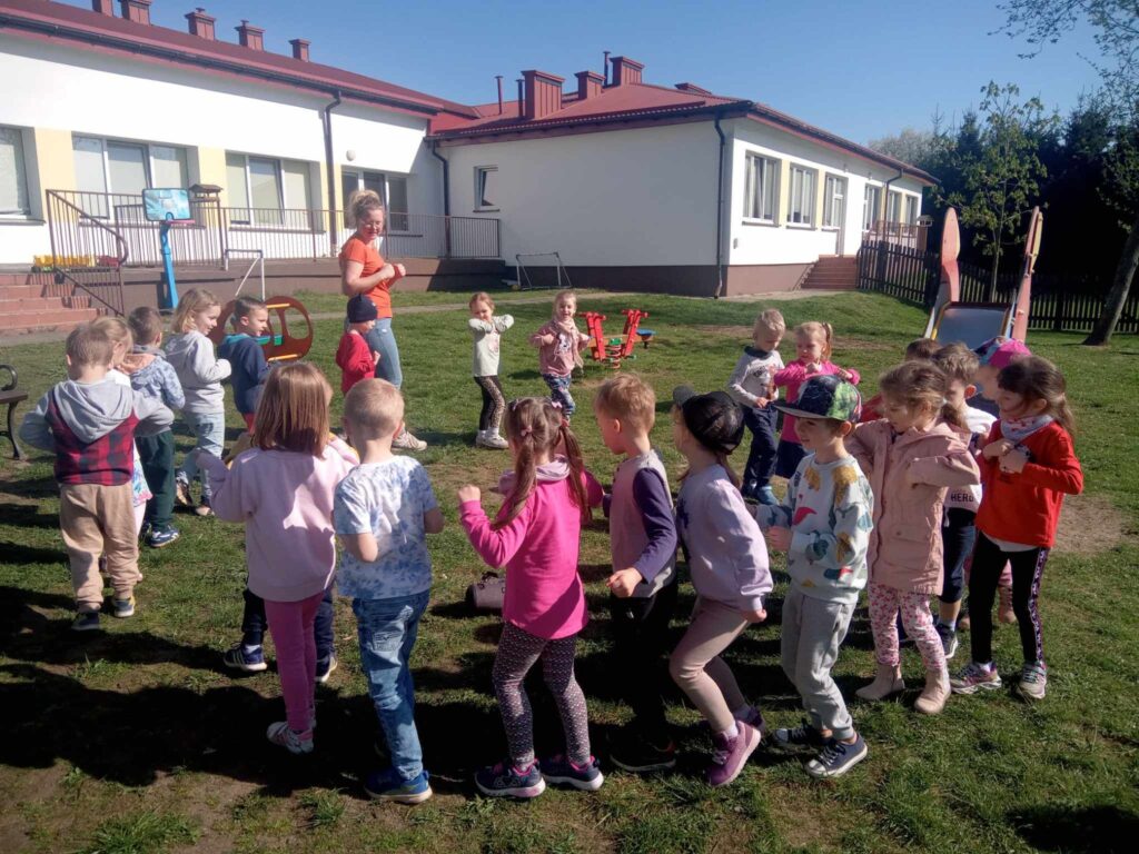 Zdjęcie przedstawia panią oraz grupę dzieci w kole w ogrodzie przedszkolnym.