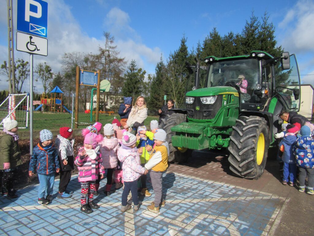 Zdjęcie przedstawia panie i pana oraz grupę dzieci. W oddali widać ciągnik, plac zabaw oraz drzewa.