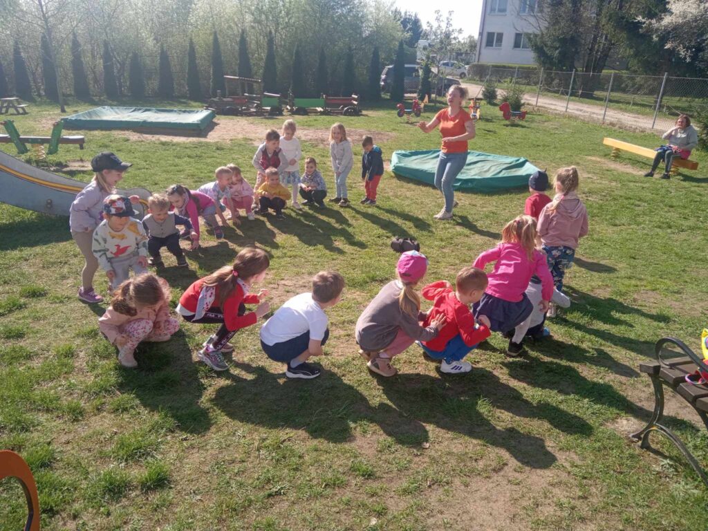 Zdjęcie przedstawia panią oraz grupę dzieci w kole w ogrodzie przedszkolnym.