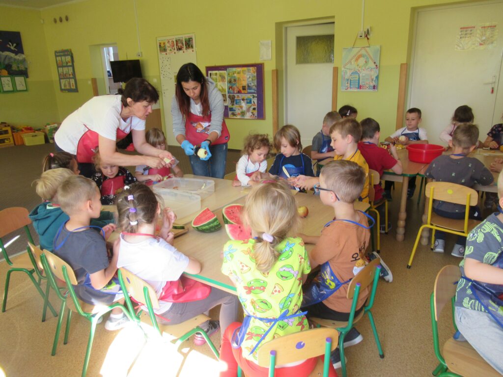 Zdjęcie przedstawia panie oraz grupę dzieci w sali przedszkolnej. Wszyscy przygotowują sałatkę.