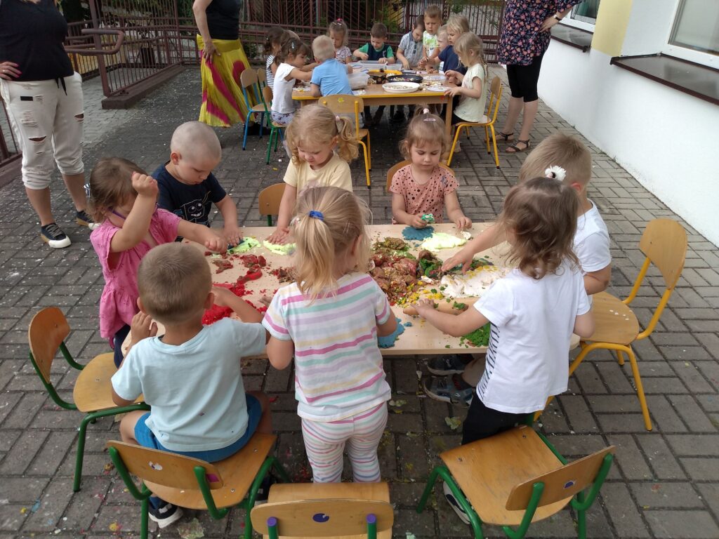 Zdjęcie przedstawia dzieci siedzące przy stolikach w ogrodzie przedszkolnym.