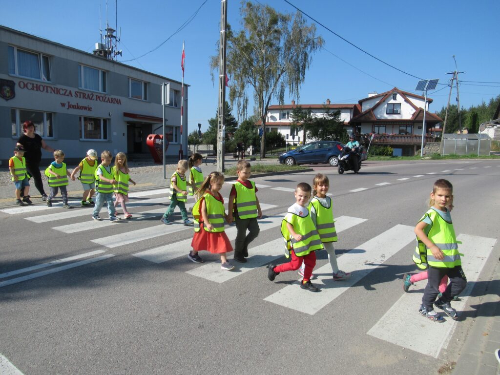 Zdjęcie przedstawia panią oraz grupę dzieci - wszyscy znajdują się na ulicy na przejściu dla pieszych. W oddali widać budynki oraz innego uczestnika ruchu drogowego.