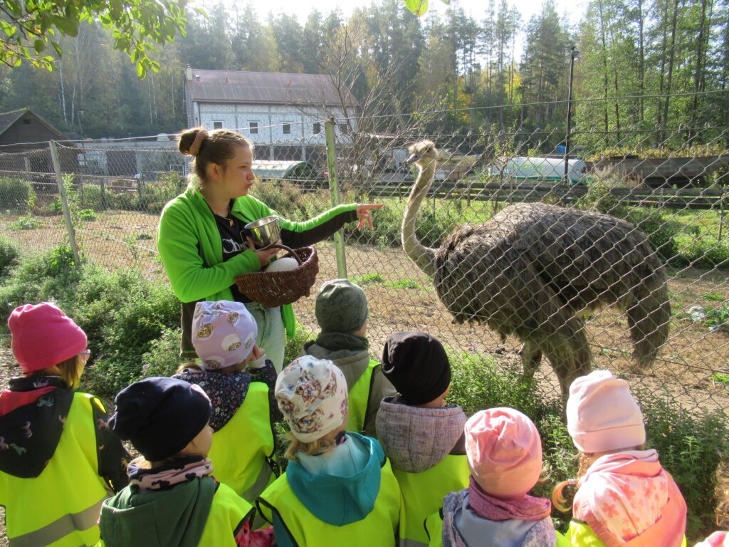 Zdjęcie przedstawia panią oraz grupę dzieci w mini zoo. W oddali widać ptaka oraz budynki