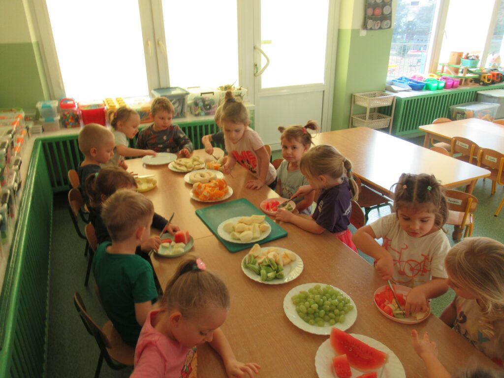 zdjęcie przedstawia zdjęcie dzieci siedzących przy stolikach w sali przedszkolnej