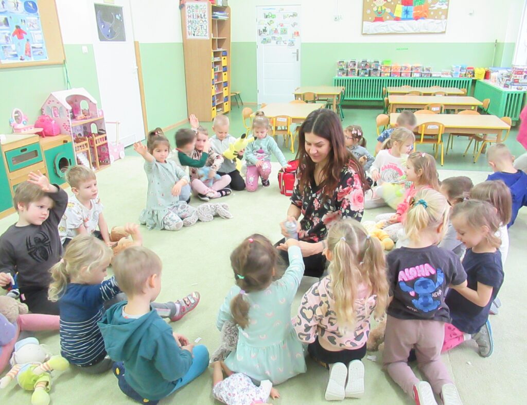 Zdjęcie przedstawia panią grupę dzieci siedzących w kole na wykładzinie w sali przedszkolnej.