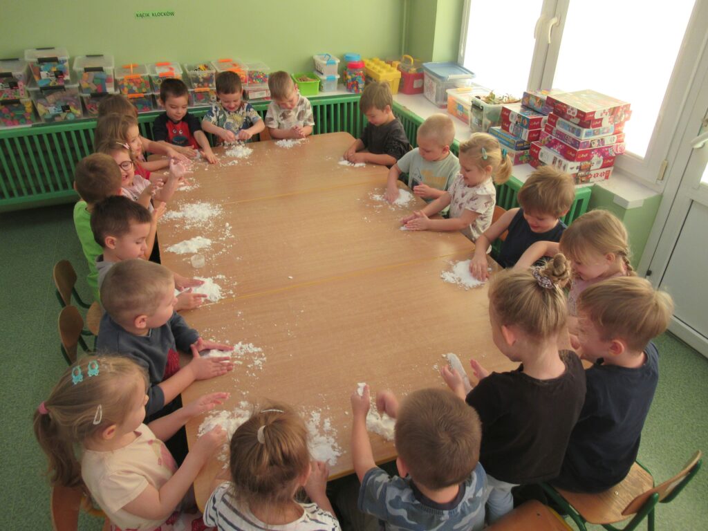 zdjęcie przedstawia zdjęcie dzieci siedzących przy stolikach w sali przedszkolnej