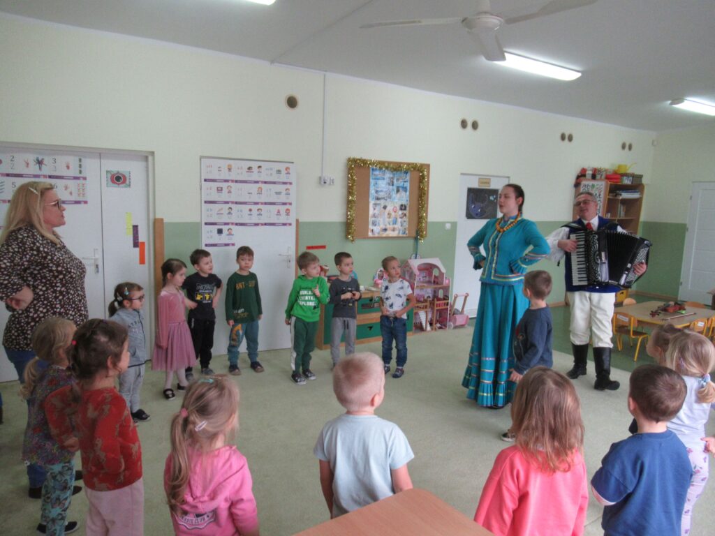 Zdjęcie przedstawia grupę tańczących ludzi w kole w sali przedszkolnej.