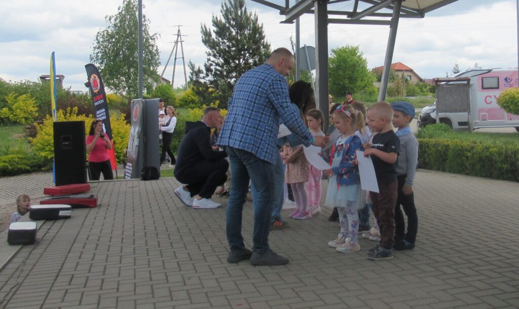 Zdjęcie przedstawia grupę ludzi na scenie, w oddali widać krzewy, dom.