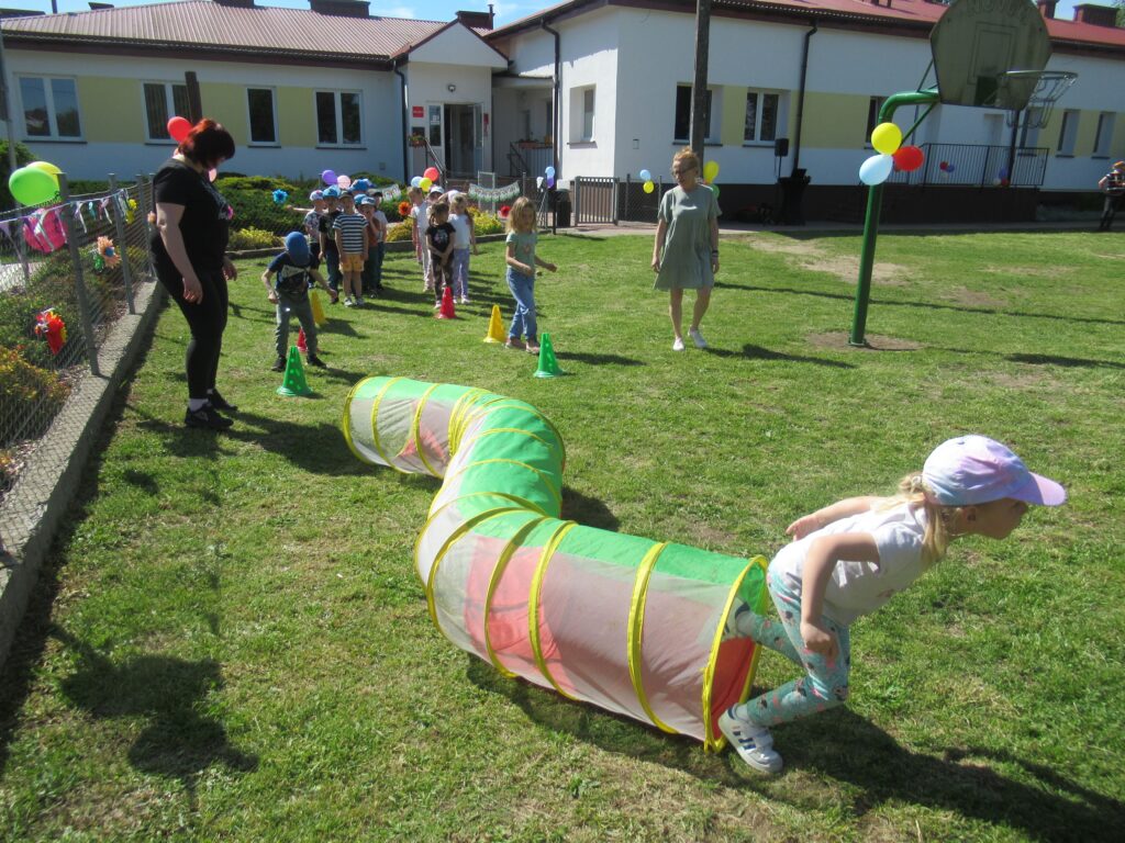 Zdjęcie przedstawia panie oraz bawiące się dzieci w ogrodzie przedszkolnym. W oddali widać budynek przedszkola.