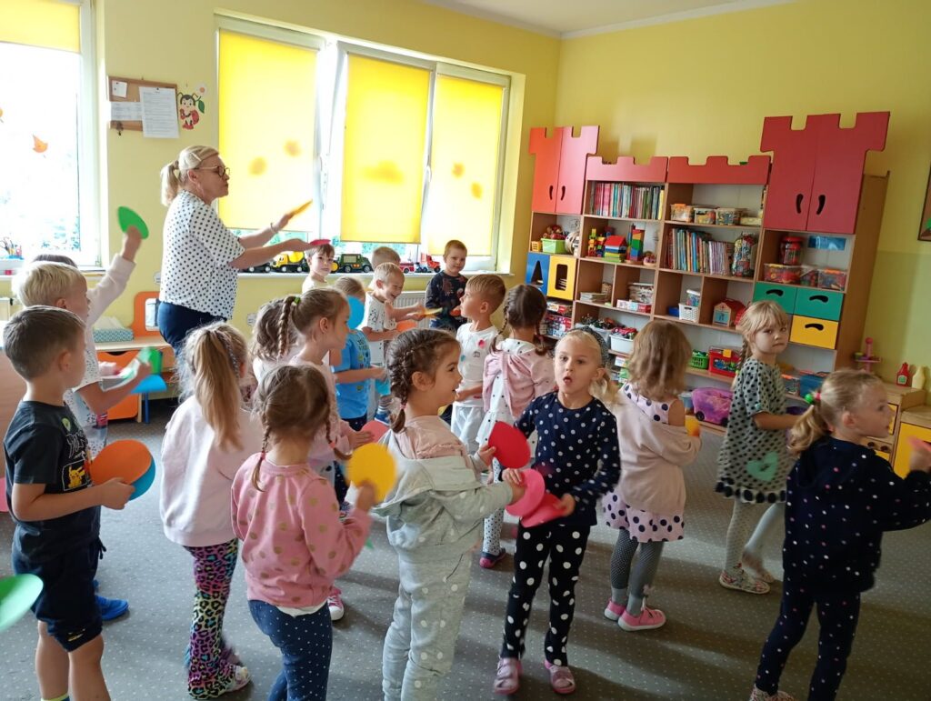 Zdjęcie przedstawia panią oraz dzieci tańczące w sali przedszkolnej.