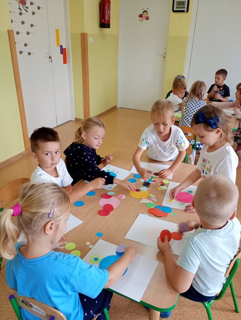 Zdjęcie przedstawia dzieci siedzące przy stolikach w sali przedszkolnej.