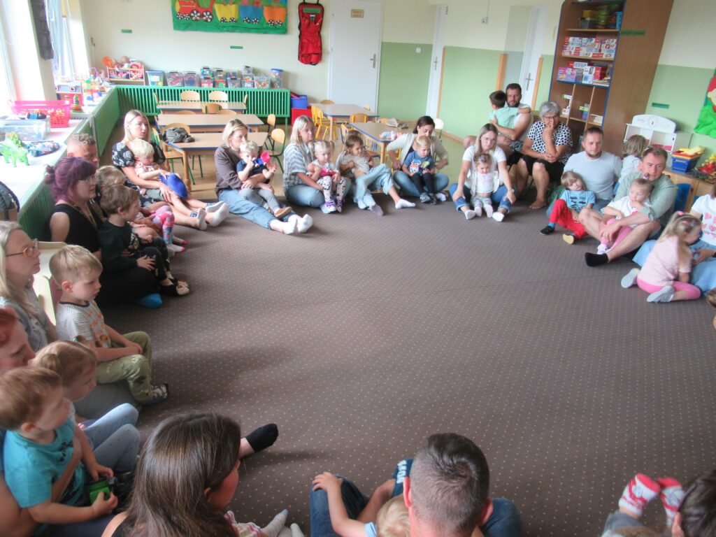 Zdjęcie przedstawia grupę ludzi w sali przedszkolnej. Wszyscy siedzą w kole na wykładzinie.