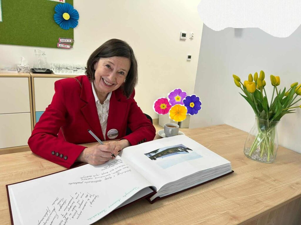 Zdjęcie przedstawia panią siedzącą przy stoliku, na stoliku leży kronika i stoją kwiaty.
