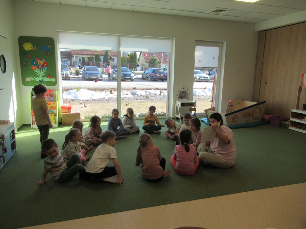 dzieci i zaproszony rodzic siedzą na dywanie, oglądają obrazki w książce