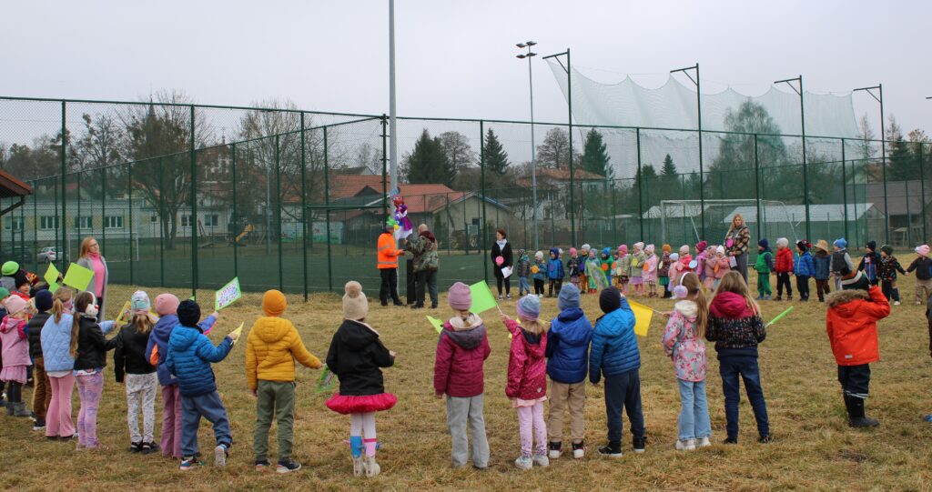 Zdjęcie przedstawia grupę ludzi na placu przedszkolnym. W oddali widać orlik i budynki.