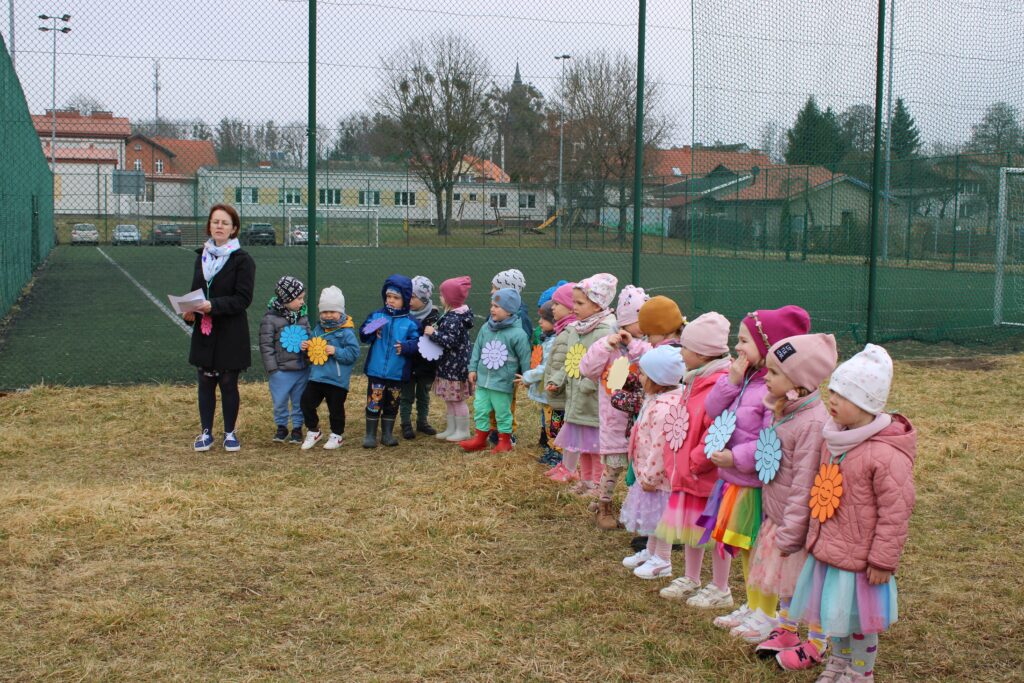 Zdjęcie przedstawia dzieci i panią na placu przedszkolnym. W oddali widać orlik, auta i budynki.