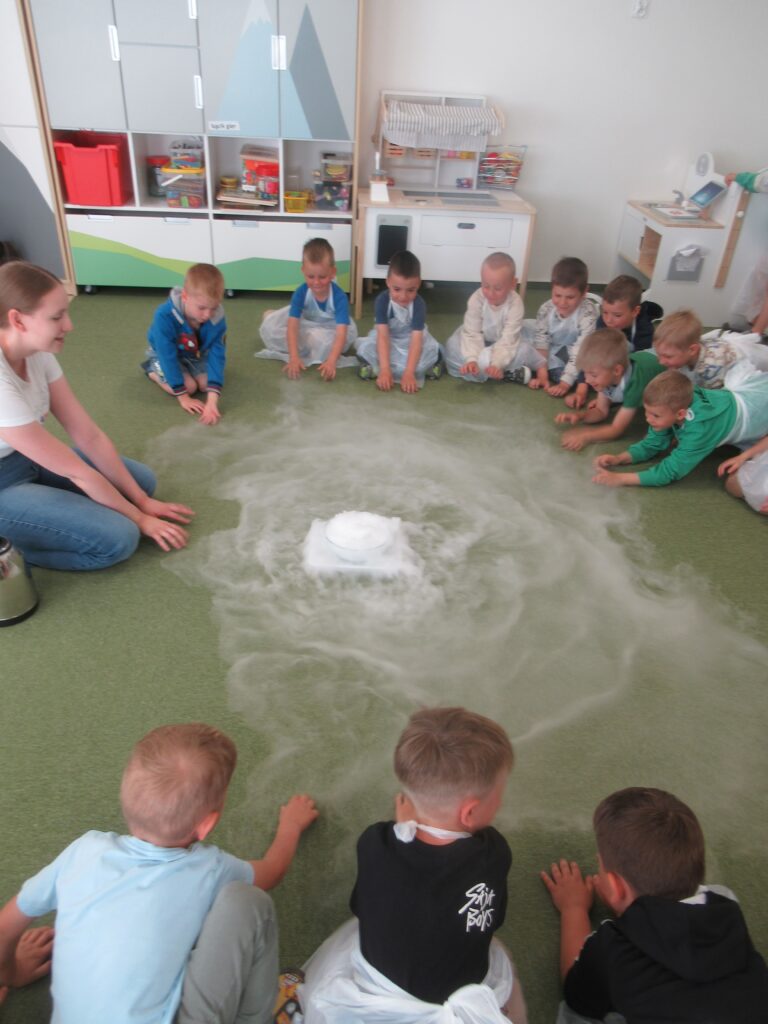 Zdjęcie przedstawia dzieci oraz panią, wszyscy siedzą w kole na dywanie.