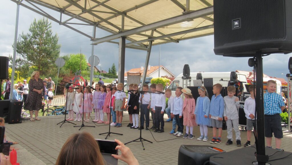 Zdjęcie przedstawia grupę ludzi na scenie. W oddali widać innych ludzi, zieleń, budynki.