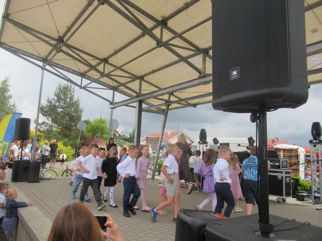 Zdjęcie przedstawia grupę ludzi na scenie. W oddali widać innych ludzi, zieleń, budynki.