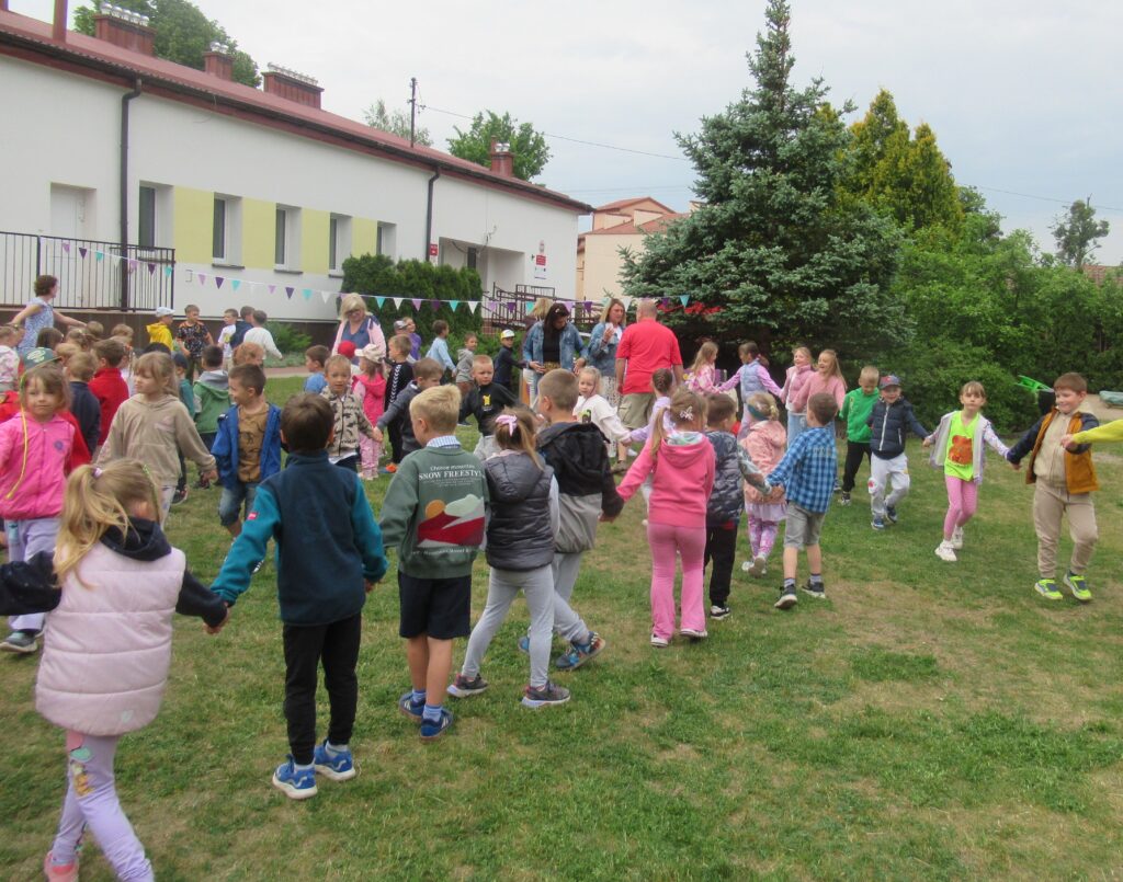 Zdjęcie przedstawia grupę bawiących się ludzi w ogrodzie przedszkolnym. W oddali widać budynek przedszkola oraz zieleń.