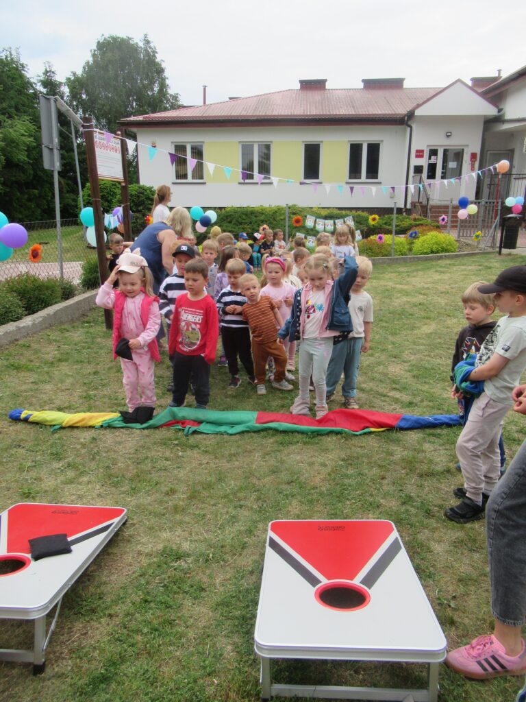 Zdjęcie przedstawia grupę dzieci w ogrodzie przedszkolnym. Z tyłu widać budynek przedszkola.