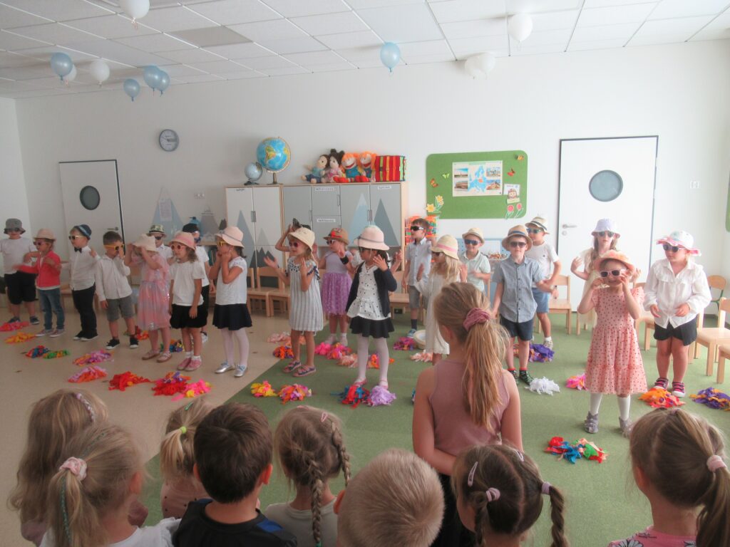 dzieci stoją w sali i śpiewają piosenkę, mają kapelusze i okulary przeciwsłoneczne